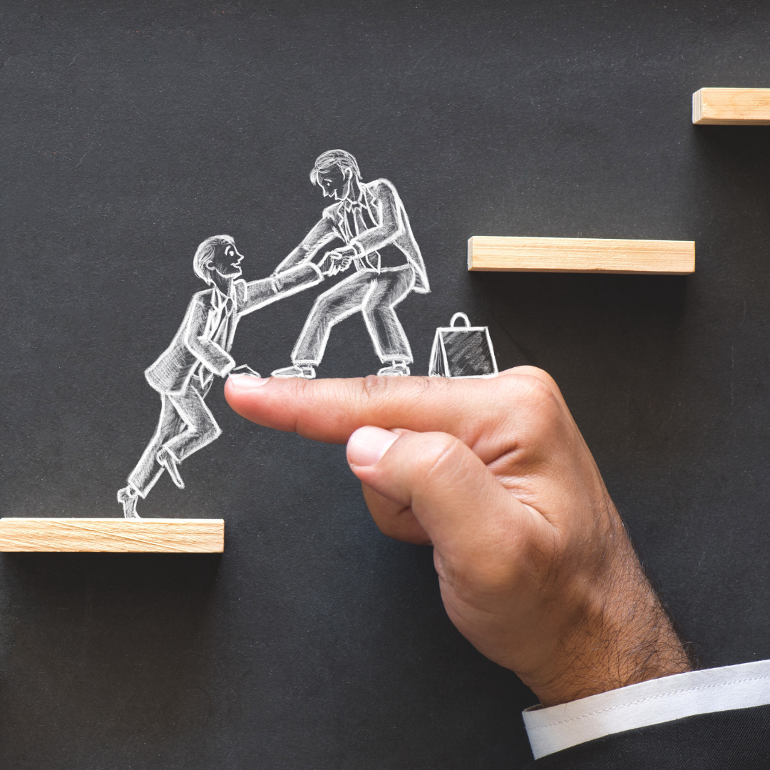 A imagem mostra um fundo escuro onde está desenhada, a giz branco, a figura de dois homens de fato: um está numa plataforma mais alta e ajuda o outro a subir, segurando-lhe a mão. Uma mão real aparece em primeiro plano, com o dedo estendido a servir de apoio entre duas pequenas prateleiras de madeira, simbolizando uma ponte. A imagem transmite a ideia de cooperação, apoio e trabalho em equipa.