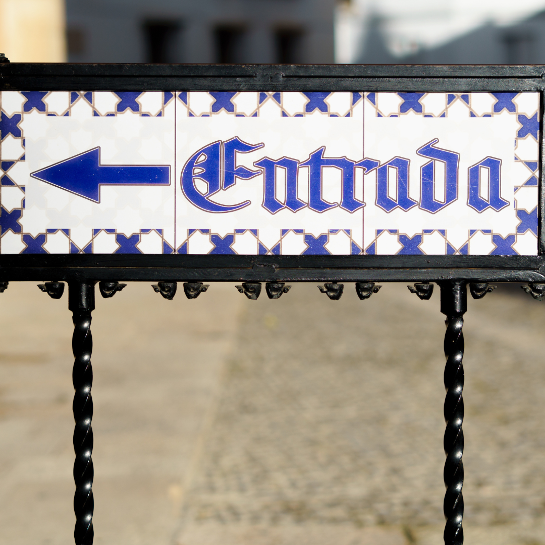 A imagem mostra uma placa decorativa em azulejos brancos e azuis com a palavra “Entrada” escrita. Ao lado do texto, há uma seta azul apontando para a esquerda, indicando a direção de entrada. A placa está fixada numa estrutura preta e o fundo parece ser uma rua pavimentada, ligeiramente desfocada.